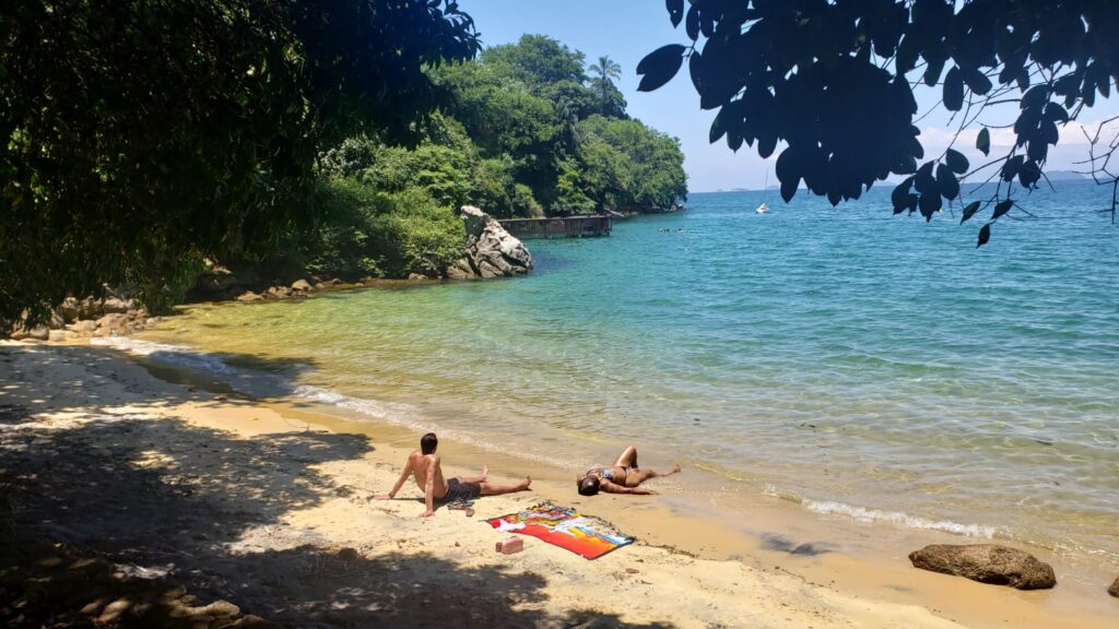 Imagem mostra duas pessoas sentadas na areia da praia da baleia